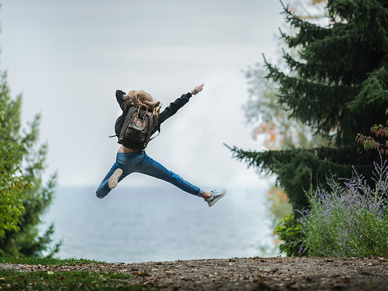 woman wearing a back pack jumping up in the air on a wooded trail